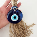 Close-up of a blue evil eye pendant, small eye beads and tan fringe held against a hand