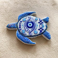 Turtle-shaped magnet with blue patterned flippers, a white shell bearing an eye motif and CRETE lettering, shown at an angle.