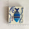 White rectangular magnet with a blue and turquoise patterned fish pointing downward, curling foliage and CRETE lettering beside ruler markings.