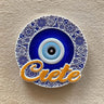 Round blue eye magnet with a blue and white floral border and orange Crete script across the bottom.