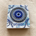 White rectangular magnet with a raised blue eye, Greek key and wave borders, leafy details and GREECE lettering.