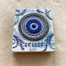 White rectangular magnet with a raised blue eye, Greek key and wave borders, leafy details and GREECE lettering.