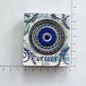 White rectangular magnet with a circular blue eye, Greek key border, leafy details and GREECE lettering beside ruler markings.