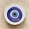 Round blue and white eye magnet with a black center, subtle wavy border and Greece lettering.