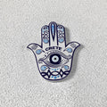 White Hamsa hand magnet with a blue eye on the palm, scrolling patterns, turquoise accents and CRETE lettering.