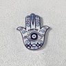 White Hamsa hand magnet with a blue eye on the palm, scrolling patterns, turquoise accents and CRETE lettering.