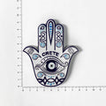 White Hamsa hand magnet with blue eye and scroll patterns, turquoise accents and CRETE lettering beside ruler markings.