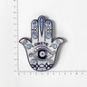 White Hamsa hand magnet with blue eye and scroll patterns, turquoise accents and CRETE lettering beside ruler markings.
