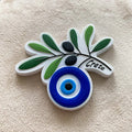 Olive branch magnet with green leaves, black olives, a blue and white eye and Crete lettering, shown at an angle.