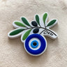Olive branch magnet with green leaves, black olives, a blue and white eye and Crete lettering, shown at an angle.