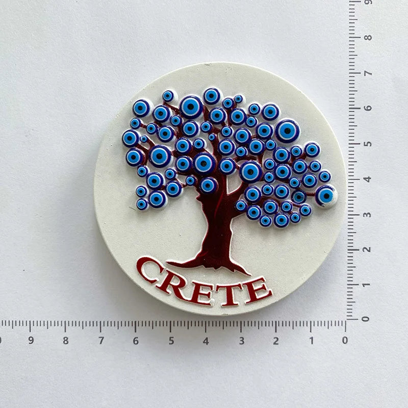 Round white magnet with a dark red tree, blue eye motifs and CRETE lettering beside horizontal and vertical ruler markings.