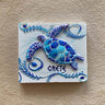 Blue and turquoise turtle on a white rectangular magnet with curling leafy borders and CRETE lettering.