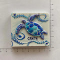 White rectangular magnet with a blue and turquoise turtle, curling leafy borders and CRETE lettering beside ruler markings.