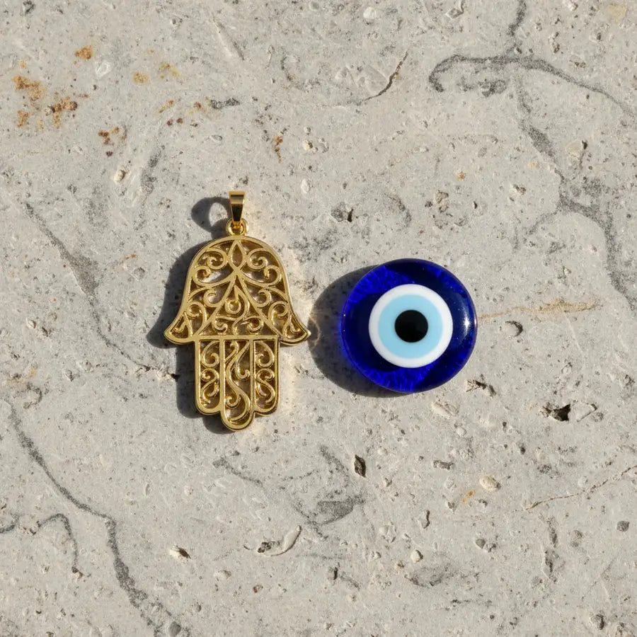 Gold Hamsa pendant and blue glass Evil Eye bead resting side by side on a light natural stone surface.
