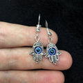 Silver Hamsa hand earrings with blue eyes held between fingers against a dark background