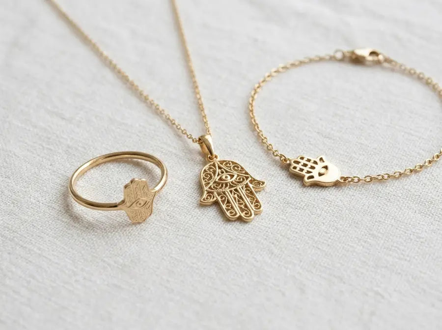 Flat-lay of a Hamsa necklace pendant, a Hamsa bracelet charm, and a Hamsa engraved ring on soft fabric