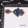 Single Blue Hamsa earring laid sideways beside horizontal and vertical ruler scales, with a printed weight annotation.