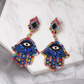 Blue Hamsa earrings at an angle on a white slab, with multicolored borders and visible posts behind the decorated tops.