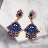 Blue Hamsa earrings at an angle on a white slab, with multicolored borders and visible posts behind the decorated tops.