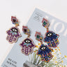 Purple and Blue Hamsa earring pairs arranged on a printed page, with diamond-shaped tops and decorated hand drops.