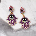 Purple Hamsa earrings at an angle, showing iridescent borders, leaf-shaped accents and short gold tone connecting links.