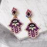Purple Hamsa earrings at an angle, showing iridescent borders, leaf-shaped accents and short gold tone connecting links.