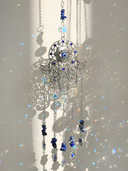 Silver tone Hamsa suncatcher with a blue central eye, clear faceted discs, dark blue beads and small hand charms.