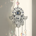 Close view of a silver tone Hamsa hand with curling open scrollwork, small blue accents and a blue evil eye in the palm.