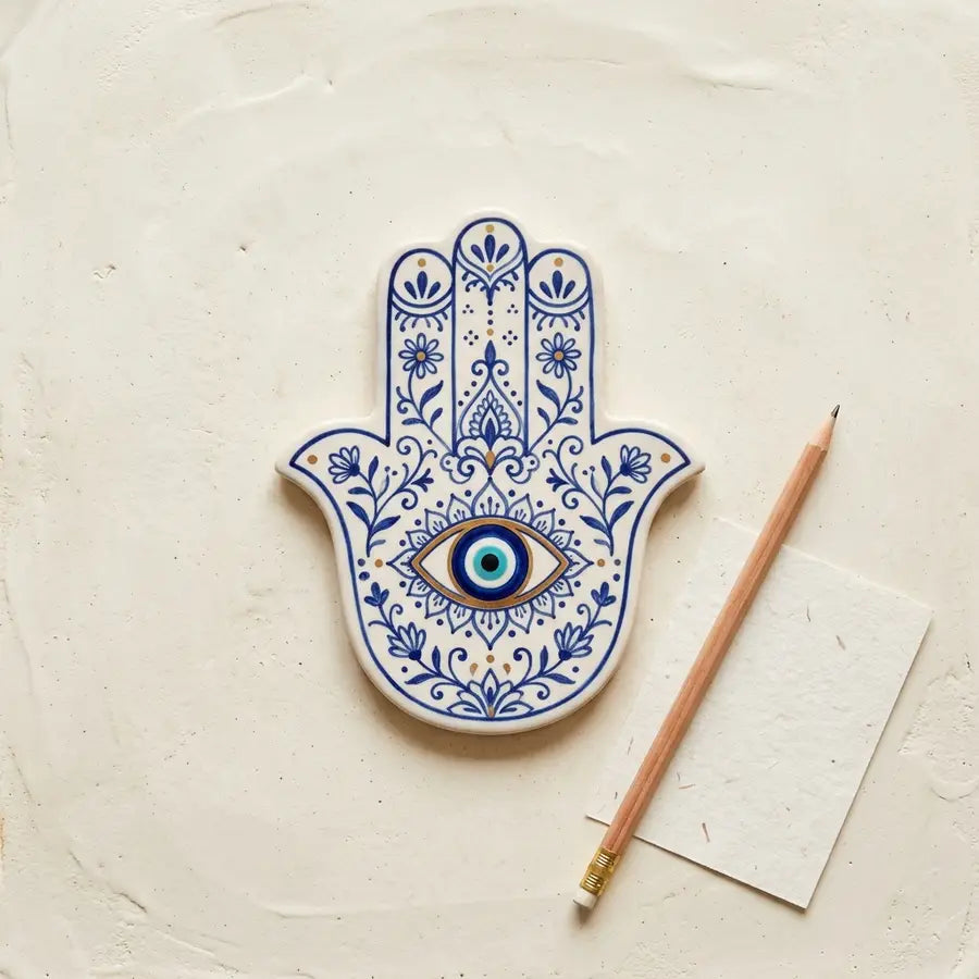 Blue floral ceramic Hamsa tile with a central eye lies beside a pencil and blank paper.