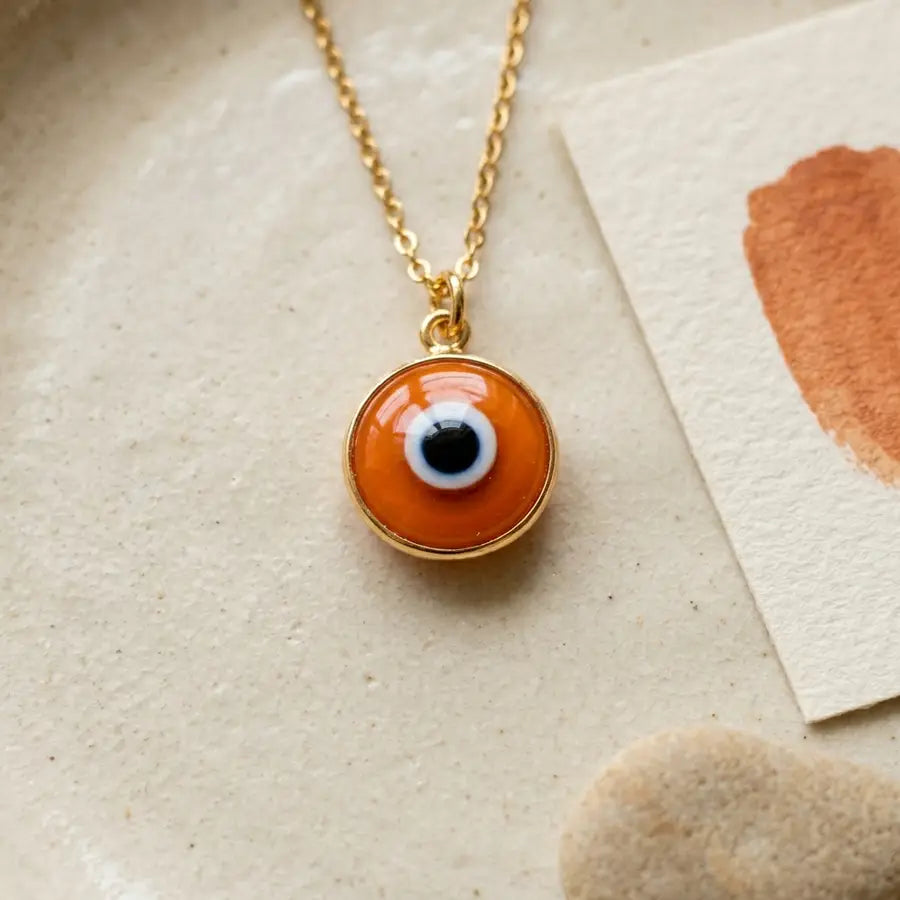 Close orange evil eye charm on a cream dish with a gold chain, terracotta brushstroke card, and smooth pebble beside it.