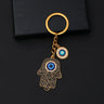 Black scroll Hamsa keychain with gold tone outlines, a blue palm eye and a separate round eye charm on a black background.