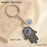 Black scroll Hamsa keychain with silver tone outlines and blue eye charms, shown with printed size and weight labels.