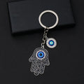 Black scroll Hamsa keychain with silver tone outlines, a blue palm eye and a separate round eye charm on a black background.