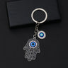 Black scroll Hamsa keychain with silver tone outlines, a blue palm eye and a separate round eye charm on a black background.