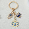 Gold tone keychain with a blue elephant, hamsa hand and textured round eye pendant, shown upright on a white background.