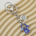 Blue hamsa keychain with silver tone ring and clasp, multicolored hand detail and round eye charm, angled across a beige ridged background.