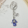 Blue hamsa keychain with silver tone details, multicolored accents around a smaller hand, and a round eye-and-ray charm, shown vertically.