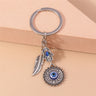 Silver tone Hamsa medallion keychain against a peach block, with blue eye details and a feather hanging to the left.