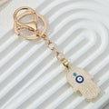 Pale pink hamsa keychain with gold tone ring and clasp, blue and turquoise eye motifs, angled across a white ridged background.
