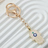 Pale pink hamsa keychain with gold tone ring and clasp, blue and turquoise eye motifs, angled across a white ridged background.