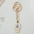 Pale pink hamsa keychain with gold tone line patterns, two eye motifs, a circular key ring and lobster-style clasp, shown vertically.