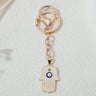 Pale pink hamsa keychain with gold tone line patterns, two eye motifs, a circular key ring and lobster-style clasp, shown vertically.