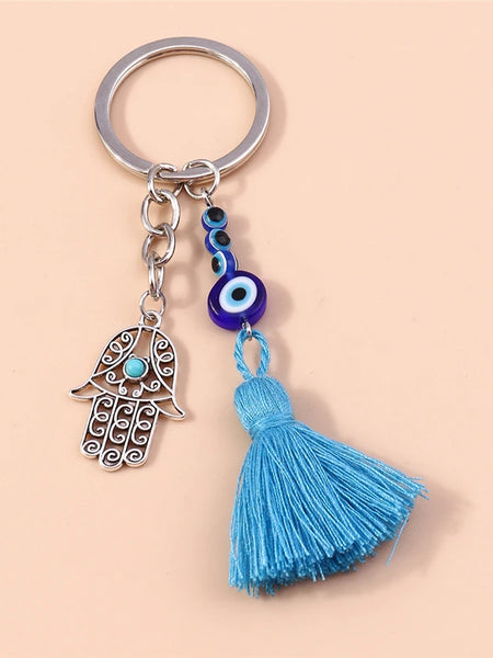 Silver tone keychain with an open scroll-patterned Hamsa hand, blue eye beads and a blue tassel on a peach background.