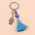 Silver tone keychain with an open scroll-patterned Hamsa hand, blue eye beads and a blue tassel on a peach background.