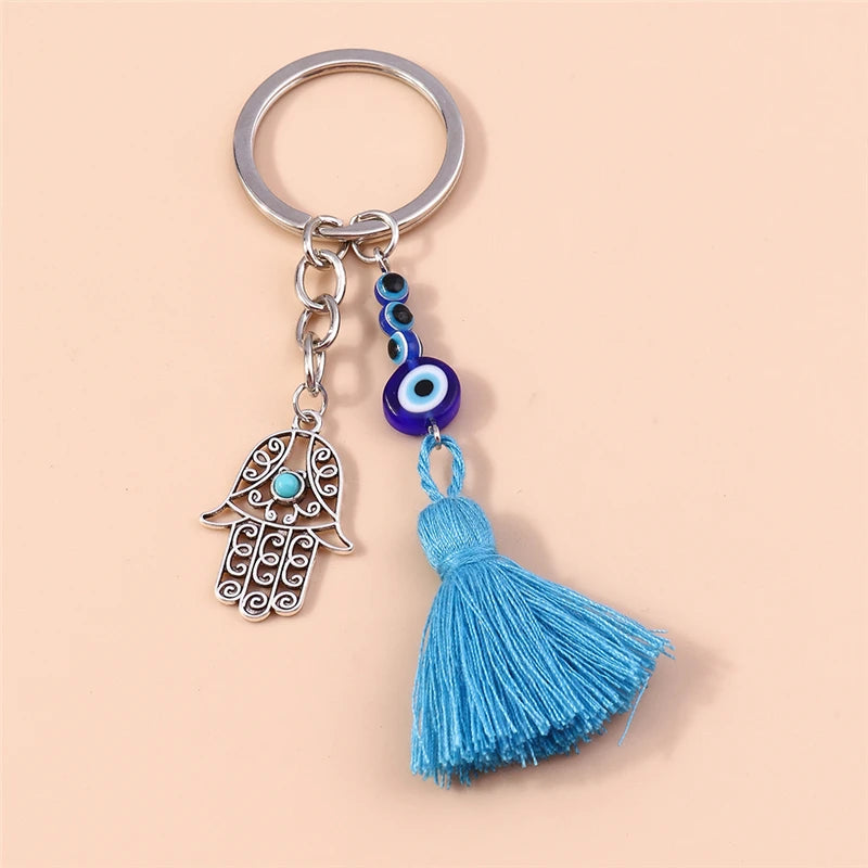 Silver tone keychain with an open scroll-patterned Hamsa hand, blue eye beads and a blue tassel on a peach background.
