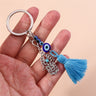 Blue Hamsa tassel keychain held across an open hand, showing the scroll-patterned charm and eye beads.