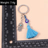 Blue Hamsa tassel keychain beside an L-shaped ruler, with a weight label in the upper left corner.