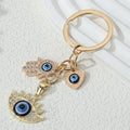 Gold tone heart cluster keychain with blue eyes on the heart, hamsa hand and pointed-lash pendant, arranged diagonally with the ring at the upper right.