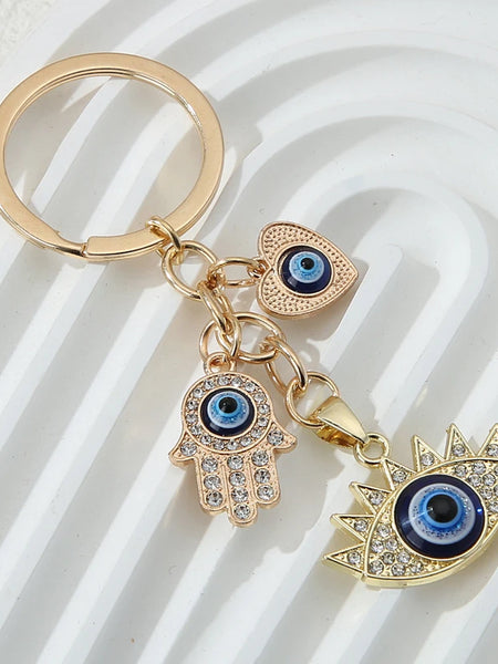 Gold tone keychain with blue eye details on heart, hamsa hand and pointed-lash eye charms, arranged on a white ridged background.