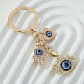 Gold tone keychain with blue eye details on heart, hamsa hand and pointed-lash eye charms, arranged on a white ridged background.